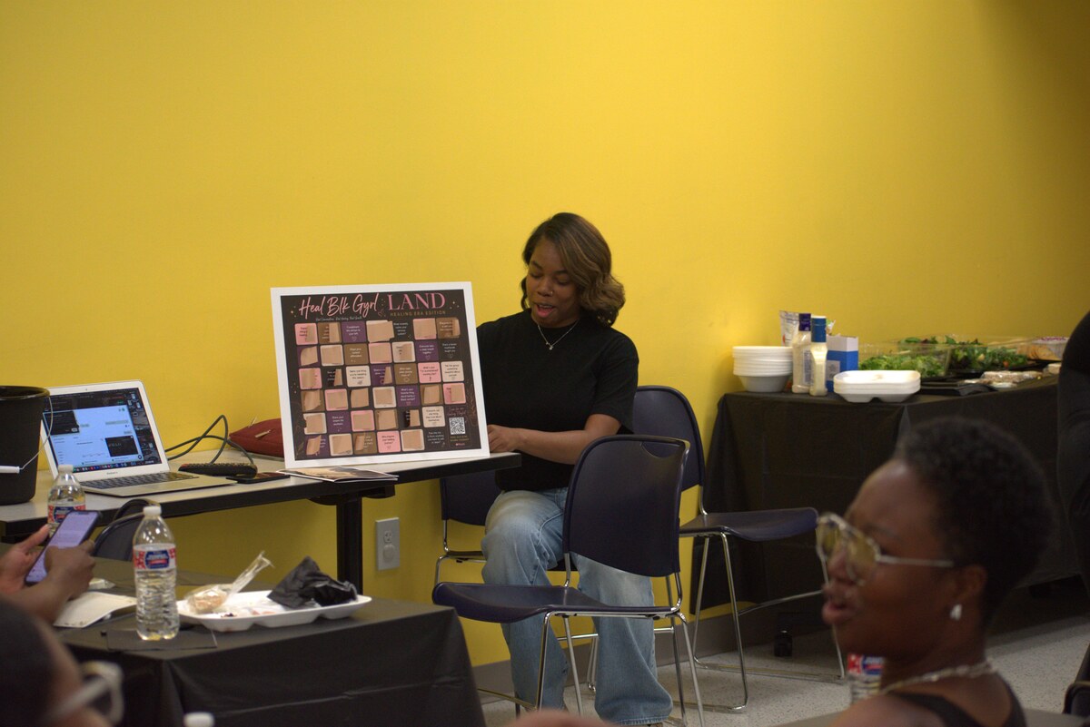 Founder Cierra Smith seated beside a Heal Blk Gyrl Land healing-era board, speaking to the room